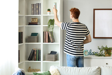 Young redhead man taking book from shelf at home, back view