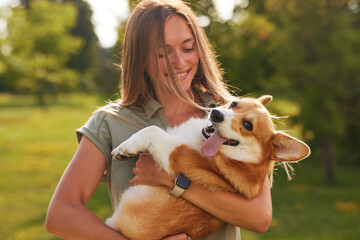 a young girl holds a cheerful and funny Welsh Corgi in her arms in a park in sunny weather, the...