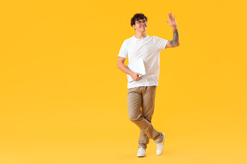 Male programmer with laptop waving hand on yellow background
