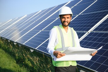 Portrait of Young indian male engineer standing near solar panels, with clear blue sky background,...