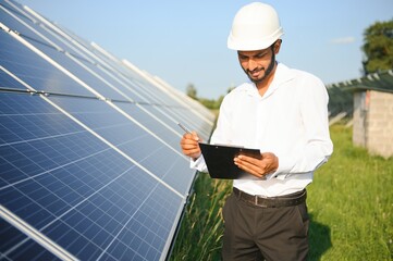 An Indian male engineer in a green vest is working on a field of solar panels. The concept of renewable energy.
