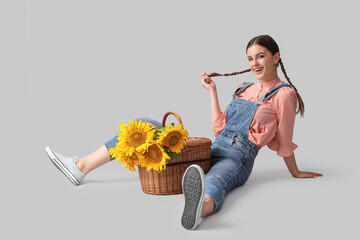 Happy young woman with wicker basket full of beautiful sunflowers sitting on grey background
