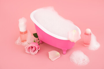 Fototapeta na wymiar Small bathtub with foam, bottles of cosmetic products and rose flower on pink background