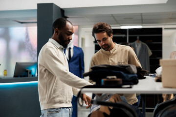 Fototapeta na wymiar Clothing store shopper and assistant having conversation about fashion trends while exploring apparel rack. African american man asking boutique worker for help in choosing outfit