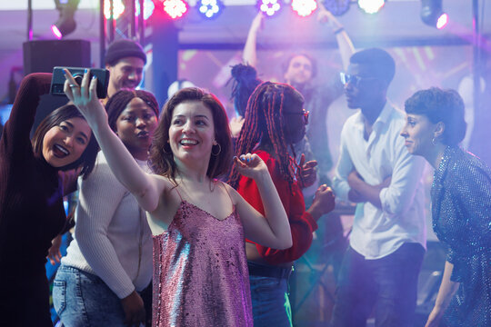 Smiling Woman Taking Selfie While Dancing With Friends At Nightclub Discotheque. Happy Girl Holding Mobile Phone And Posing For Photo On Club Dancefloor Illuminated With Stage Spotlights