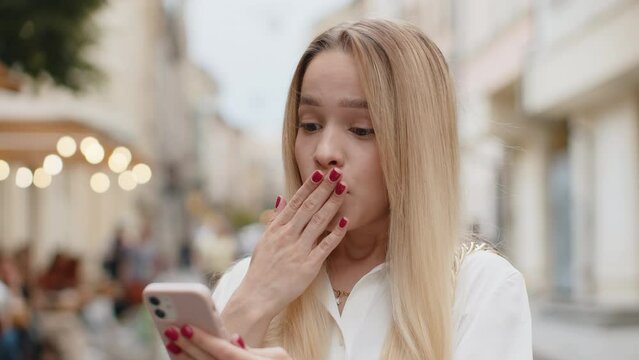 Oh my God Wow. Excited happy joyful winner blonde young woman use smartphone typing browsing celebrating win good message news. Teenager girl with mobile phone walking in urban city street outdoors