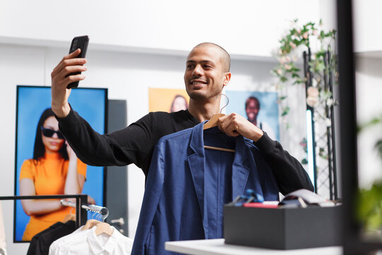 Smiling arab man holding hanger with stylish jacket while taking selfie using smartphone front camera in clothing store. Blogger showcasing apparel to social media channel followers in boutique