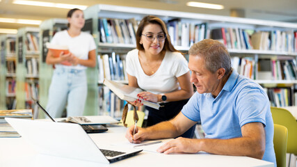 Obraz premium Concentrated mature man spending time in library, reading books and making laptop. Self-education concept