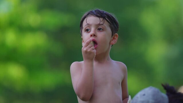 One pensive little boy standing outside shirtless after swimming at pool eating biscuit, thoughtful child with contemplative gaze drying while taking a bite of cookie