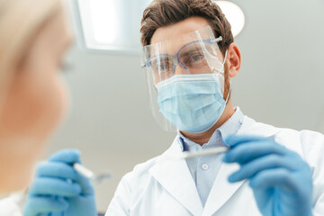 Handsome male dentist in protective mask and glasses, latex gloves checking teeth of female patient