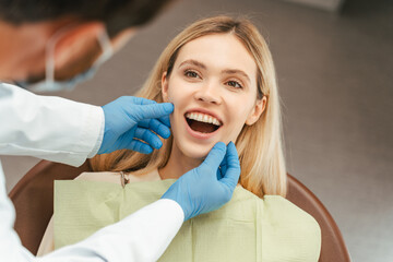 Young female patient with blond hair sitting in dental chair with open mouth, dentist checking teeth