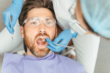 Portrait of handsome man patient sitting in dental chair wearing protective glasses with open mouth