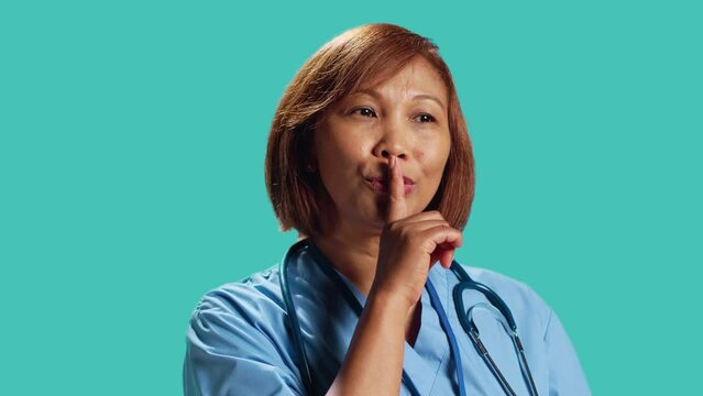 Close up shot of rigid strict BIPOC clinic manager shushing at camera, bothered by too much noise. Rigurous nurse holding quiet gesture finger to lips, isolated over studio background