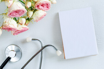 Bouquet of pink rose flowers and stethoscope with empty card notebook on a white background. National Doctor s day greeting card. Top view flat lay, copy space