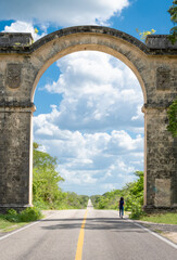 Beautiful scenery of a long, straight, empty highway, surrounded by trees, passing through a stone archway. Woman walks alone on the road