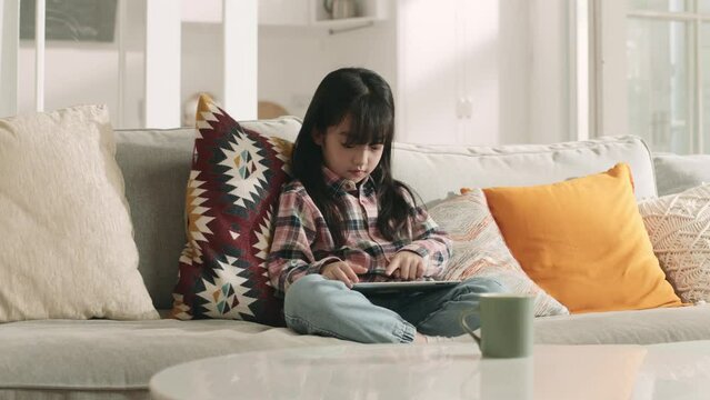seven-year-old little asian girl sitting on family couch playing computer game using digital tablet