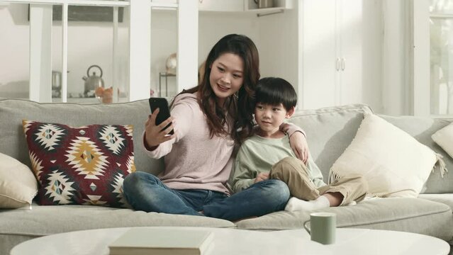 young asian mother and five years old son sitting on couch at home taking a selfie