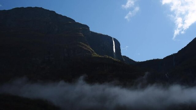 View of a dark mountain range with a sun lit waterfall  and clouds