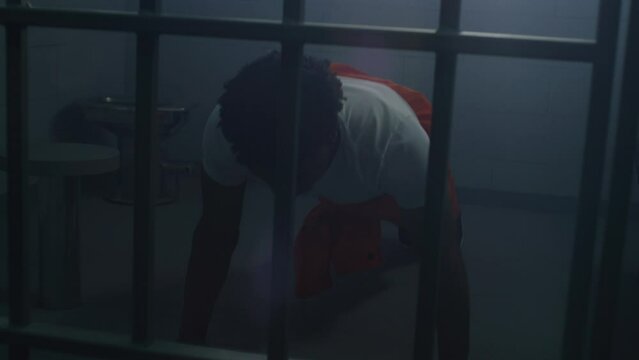 African American prisoner in orange uniform does push-ups and shadow boxing in prison cell. Inmate serves imprisonment term for crimes in jail. Criminal in detention center or correctional facility.