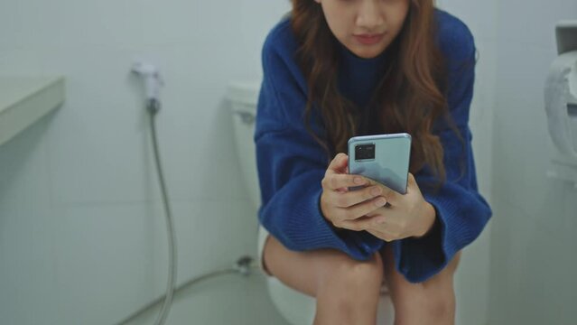 Young woman using smartphone sitting on toilet bowl at home bathroom.