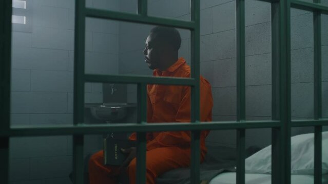 African American prisoner in orange uniform sits on the bed behind bars, reads Bible in prison cell. Criminal serves imprisonment term for crime in jail. Detention center or correctional facility.