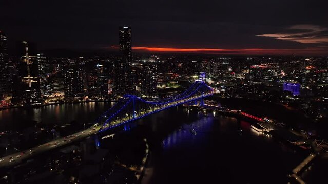 Establishing drone shot of Brisbane City's Story Bridge. Pull away night shot, Bridge lit up with Blue lights. City silhouetted over sunset red skyline.
