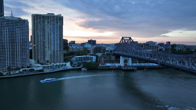 Drone establishing take off shot from above Captain Burke Park looking towards Story Bridge and Brisbane CBD. Citycat boat crossing river. Shot in evening during sunset.