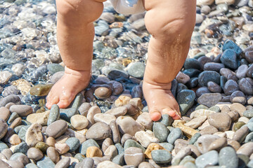 Infant boy standing on pebbles with wet feet on sunny beach. Child casting shadow explores pebbles on sunlit sea beach at seaside resort