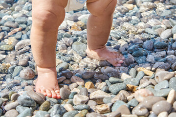 Infant boy standing on pebbles with wet feet on sunny beach. Child casting shadow explores pebbles on sunlit sea beach at seaside resort