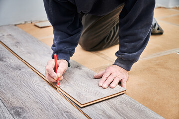Close up of male construction worker with pencil in hand drawing line on laminate wooden panel. Man preparing laminate boards for floor installation in apartment under renovation.