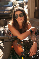 Fototapeta na wymiar Close-up portrait of a girl in sunglasses and with a tattoo on her arm, sitting on her retro motorcycle on an old cobblestone street in Europe