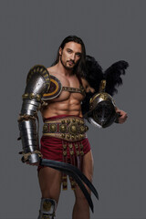 Naklejka premium Powerful and attractive gladiator with a stylish beard and luscious locks wears ornate lightweight armor, clutching a gladius and feathered helmet as he stands confidently against a grey backdrop