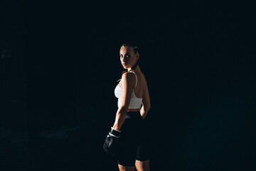 Fototapeta premium girl boxer standing in a confident pose surrounded by twilight. An expression of determination on the face, contrasting shadows, an atmosphere of intense preparation.