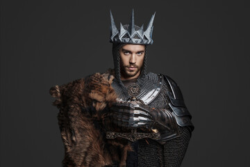 King wearing chainmail with reinforced steel plates, one shoulder covered by a fur pelt, strikes a regal pose while holding a sword against a neutral gray background in a portrait