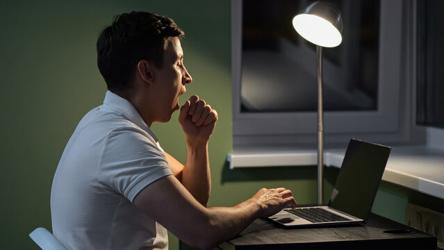 Man feels pain in eyes from load of working at laptop in semi-dark premise with floor lamp. Male worker yawns tired of overwork sitting at wooden table