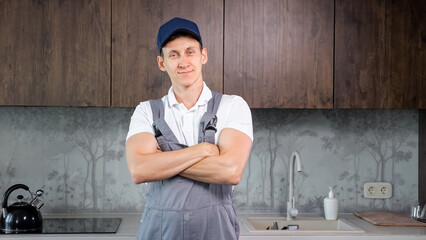 Positive male plumber in uniform stands with arms folded and smiles to camera in kitchen premise. Plumbing repair services in apartments