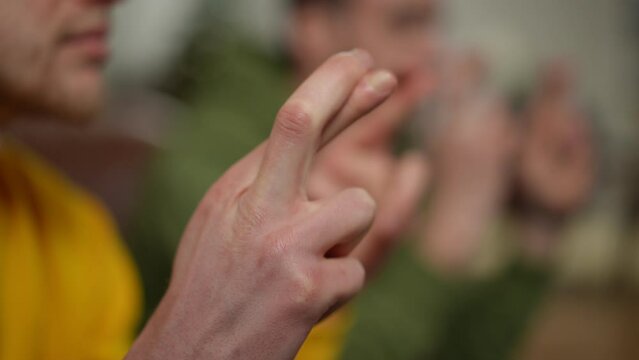 Closeup male hand with crossed fingers indoors. Unrecognizable Caucasian young man making wish sitting with blurred friend on couch at background