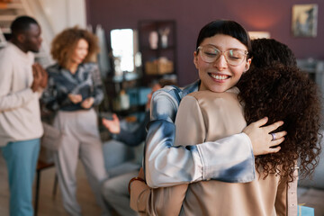 Young cheerful brunette woman in eyeglasses embracing her friend after giving her bithday present against grup of intercultural people