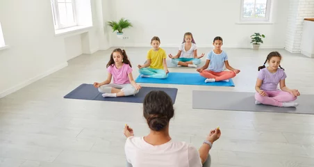 Foto auf Acrylglas Group of kids having a yoga class. Several children doing relaxing exercises. Little girls in sportswear sitting on floor mats in a lotus pose and learning to mediate with a woman coach trainer © Studio Romantic