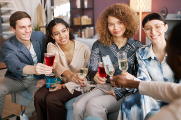 Row of happy intercultural girls and guy with glasses of alcoholic drinks clinking with that of young African American man during home party