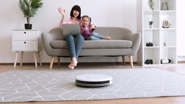 Attractive woman programing robotic vacuum via remote control while sitting behind laptop with kid on sofa. Caucasian mother and daughter watching cartoons online while tech gadget cleaning carpet.
