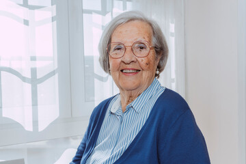 Smiling elder mature grey haired woman looking at camera, happy old lady in red eyeglasses posing at home indoor, positive single senior retired female headshot portrait.