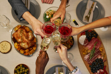 Top view of hands of young intercultural friends with alcoholic drinks clinking with flutes and wineglasses over table served for dinner