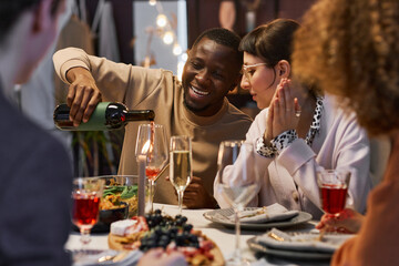 Happy young African American man pouring red wine into flute of his girlfriend while sitting next to her by served table at home party