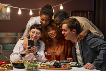Group of happy young intercultural friends communicating in video chat while looking at screen of smartphone held by smiling brunette girl