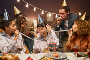 Group of intercultural young people in birthday caps blowing noise makers into ears of smiling brunette girl while sitting by served table together - obrazy, fototapety, plakaty