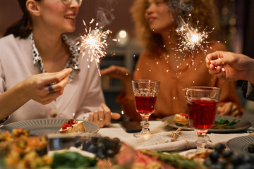 Close-up of happy young friends holding sparklers over served table with homemade food and snacks and two glasses of red wine at party