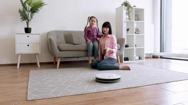 Caring woman cuddling preteen girl on sofa while sitting on floor with switched on robot vacuum cleaner. Mother-daughter duo sharing valuable moments while smart device removing dust from carpet.