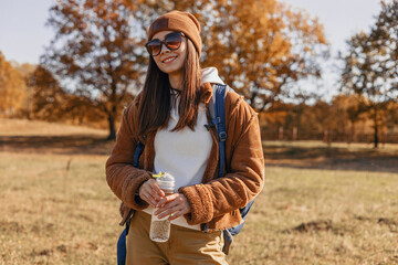 Cheerful young traveling woman with backpack and water bottle standing in nature in sunny autumn day 