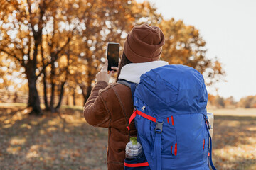 Female backpacker with smartphone taking photo in autumn forest during travel 